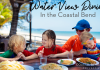 Best Waterfront Restaurants in Corpus Christi, Port Aransas & Rockport Image: A mom wearing an orange shirt and blue baseball cap, is eating lunch with three children. From left to right, there is a blond haired boy wearing a green shirt, he is talked to a brunette little girl, wearing a red shirt. Sitting on mom's lap is a child wearing goggles and a rainbow striped sun shirt. They are eating lunch with the ocean behind them.
