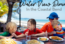 Image: A mom wearing an orange shirt and blue baseball cap, is eating lunch with three children. From left to right, there is a blond haired boy wearing a green shirt, he is talked to a brunette little girl, wearing a red shirt. Sitting on mom's lap is a child wearing goggles and a rainbow striped sun shirt. They are eating lunch with the ocean behind them.